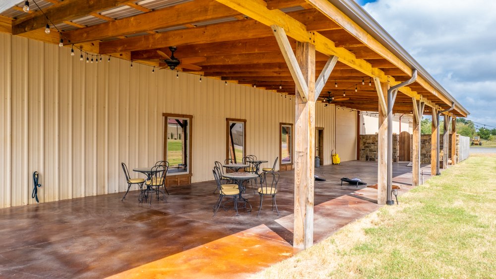 Outdoor covered patio area with wooden beams, string lights, a ceiling fan, and outdoor furniture, including a table with chairs, attached to a building with beige siding and nearby grassy yard.