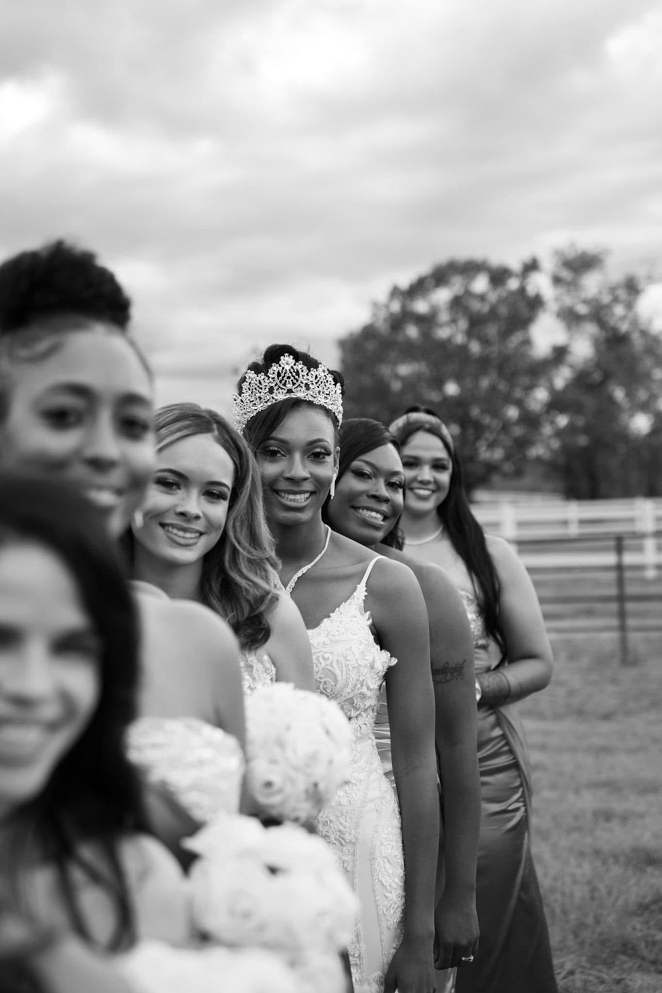 Group of women, one wearing a crown, standing in a line outdoors with trees and cloudy sky in the background.