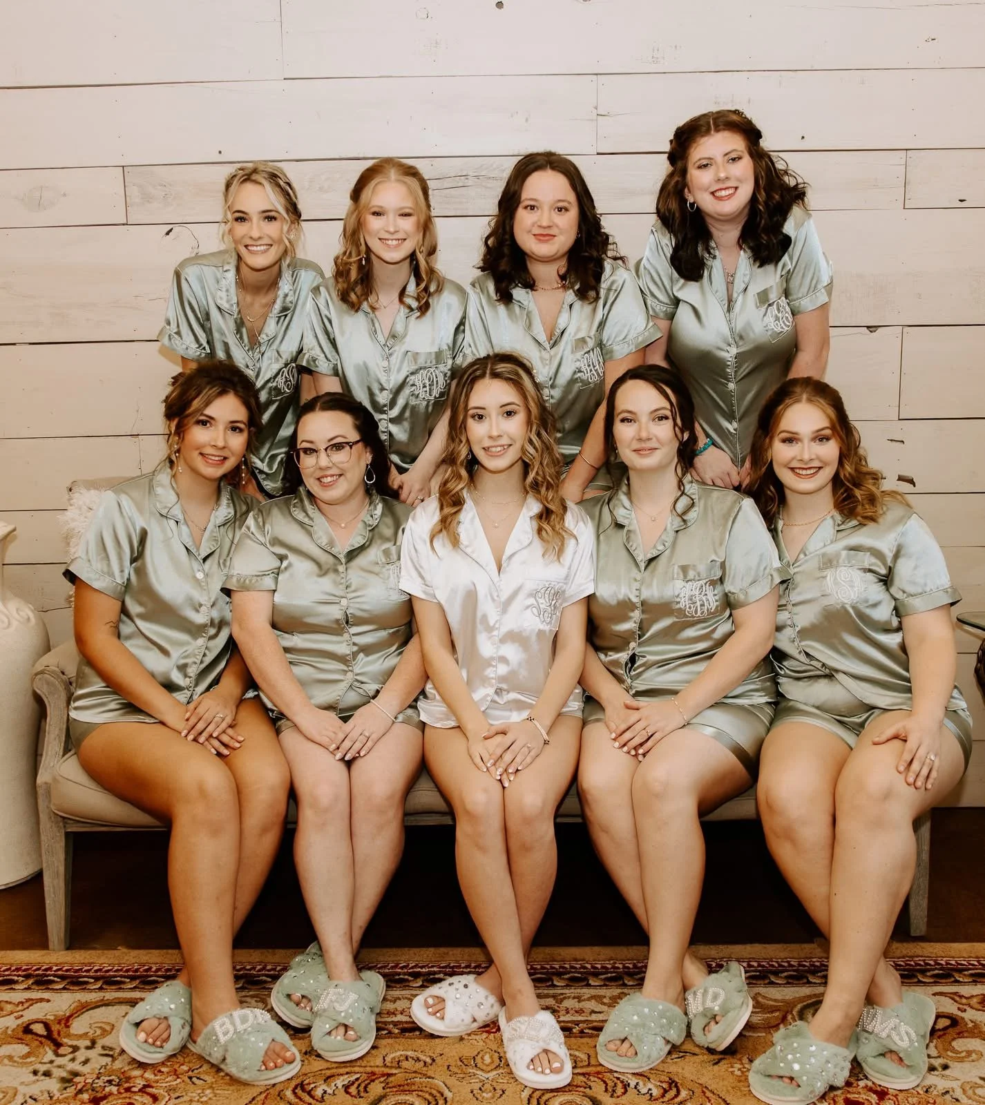 Group of nine women in satin pajamas sitting and standing on a sofa, smiling, with a wooden wall background, in a bridal or sleepover setting.
