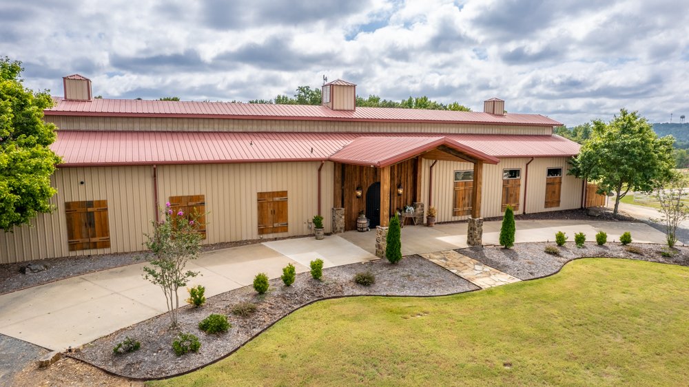 A large barn-style building with a red metal roof, wooden accents, and shudders over the windows, situated in a landscaped yard with green grass, trees, and a concrete driveway.