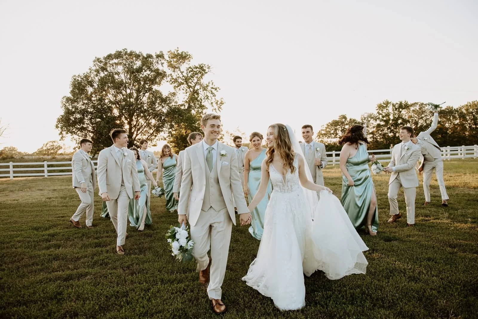A wedding party walking across a grassy field with a white fence and trees in the background during sunset, including a bride and groom in the center with bridesmaids and groomsmen surrounding them.