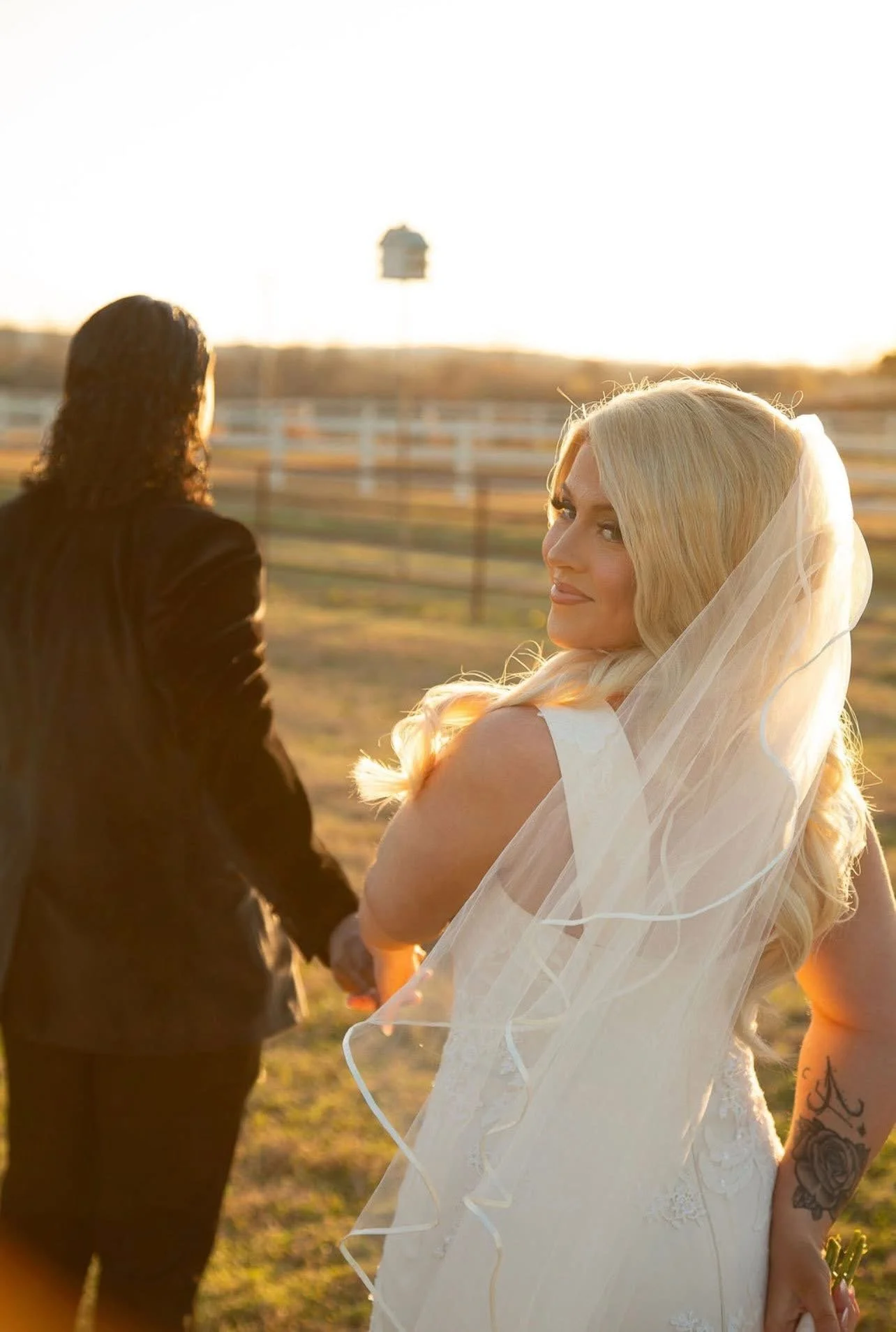 A bride and groom holding hands outdoors during sunset, with the bride smiling and wearing a wedding dress and veil, and the groom dressed in a dark suit, near a wooden fence and a field.