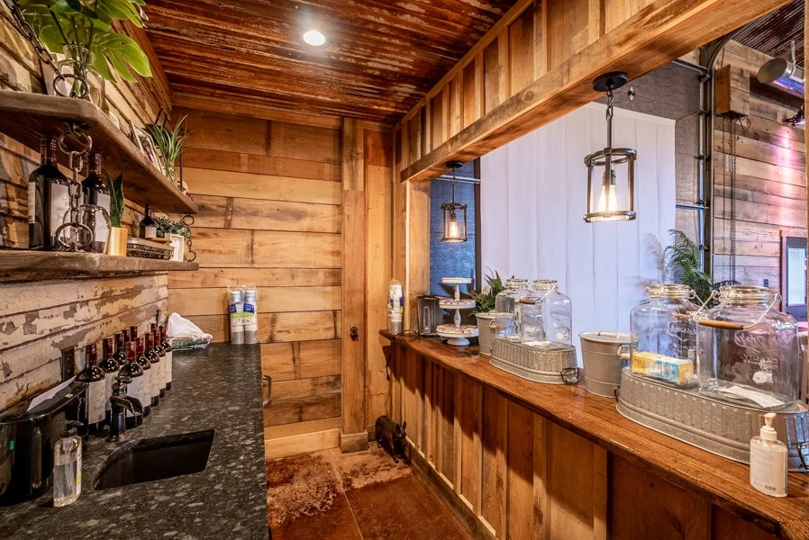 A rustic cafe with wooden walls, shelves holding decorative plants and bottles, a countertop with water bottles, and glass dispensers for drinks, illuminated by hanging lantern-style lights.