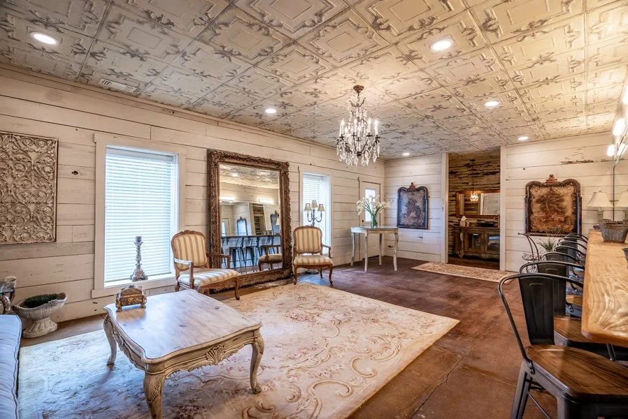 A spacious living room with white wooden walls and a decorative ceiling with chandelier, featuring vintage-style furniture, a large mirror, and a cream-colored rug.