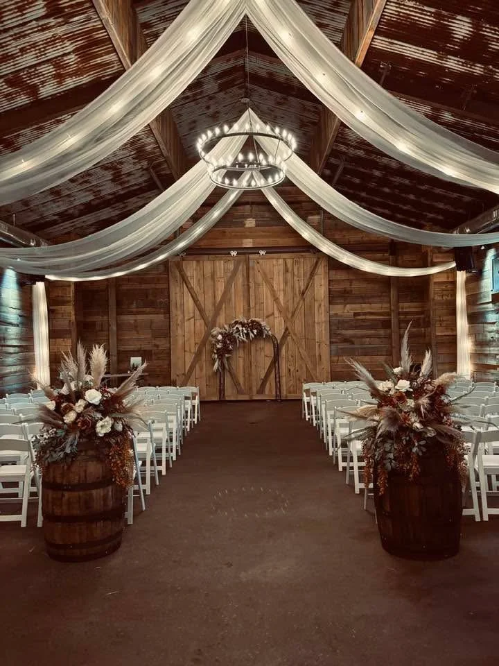 Wedding ceremony setup inside a rustic barn with white chairs, floral arrangements in wooden barrels, draped fabric on the ceiling, and a heart-shaped floral arch at the front.