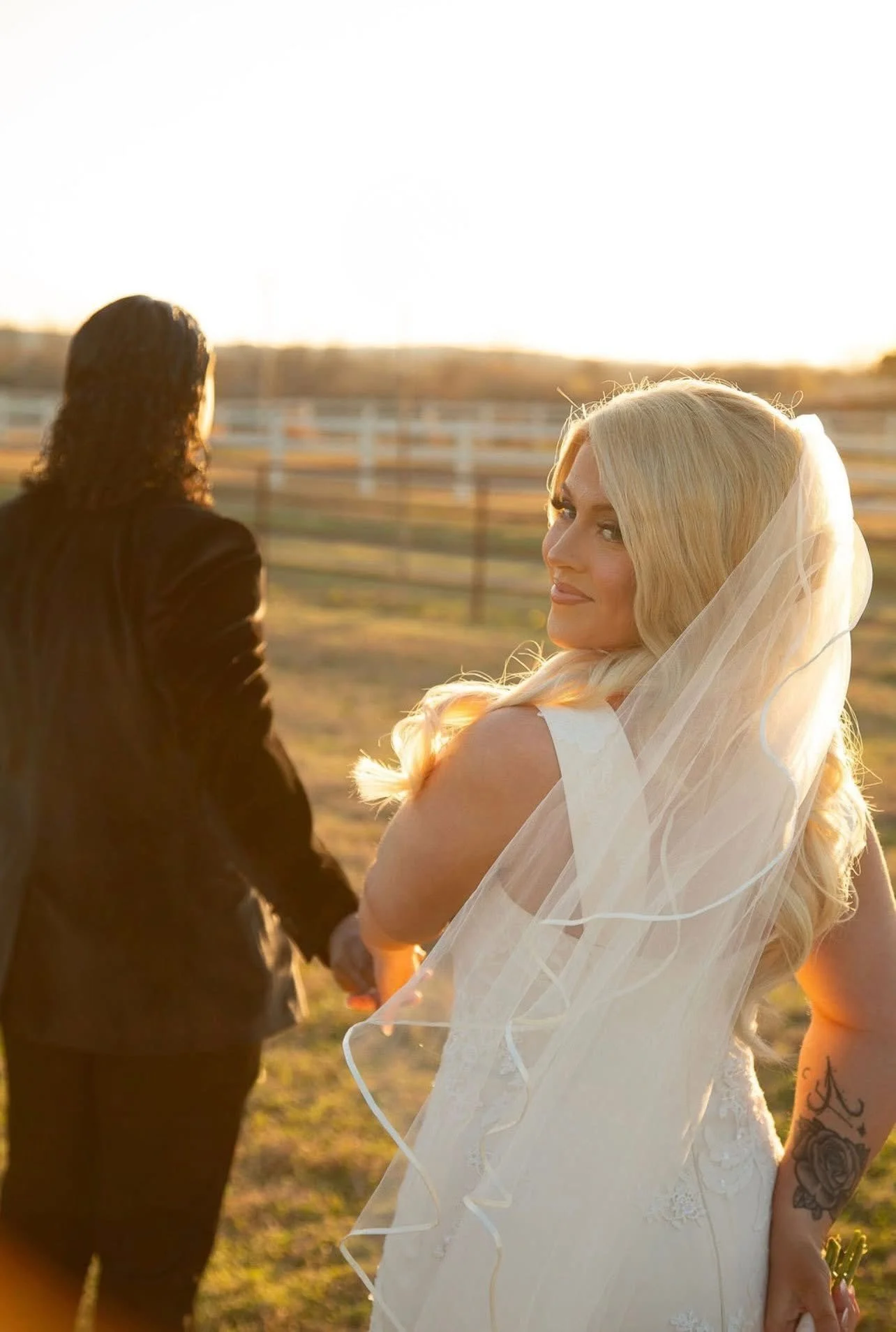 A bride with long blonde hair in a wedding dress holding hands with her partner, with a woman in a black outfit facing away, outdoors during sunset.