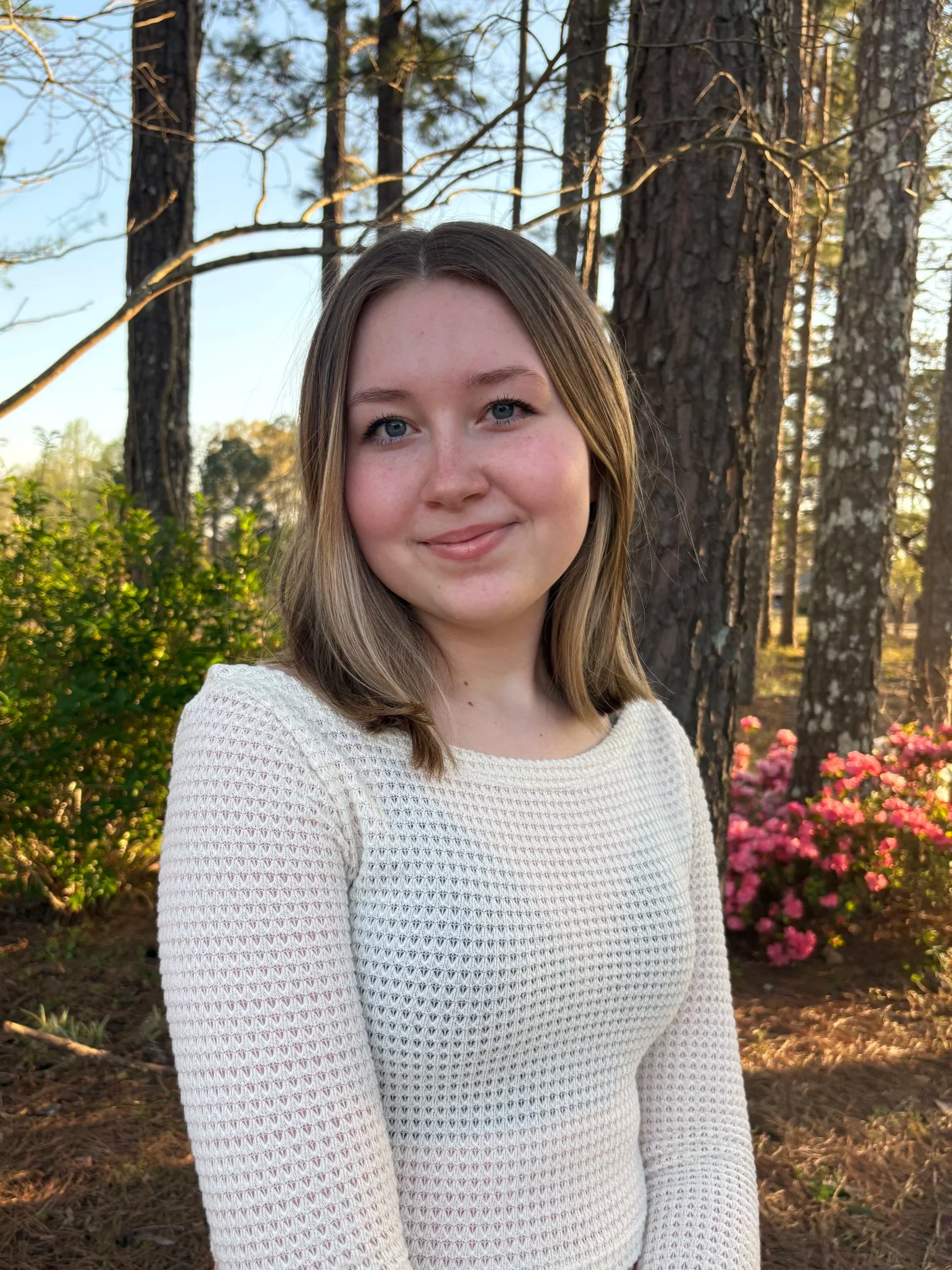 A young woman with shoulder-length brown hair and blue eyes smiling outdoors in front of tall trees and pink flowers, wearing a white knit sweater.
