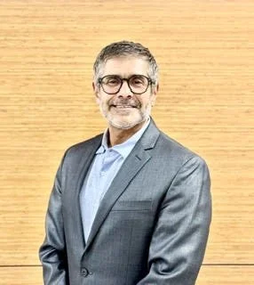 Portrait of a smiling middle-aged man with gray hair and glasses, wearing a gray suit and light blue shirt, standing in front of a wood-paneled wall.