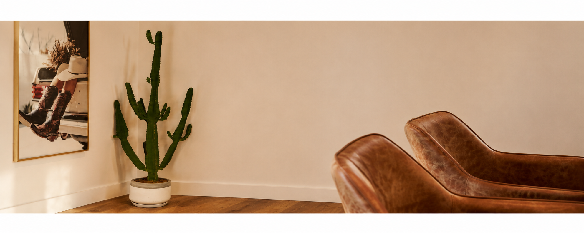 Interior room with a large cactus plant in a white pot, a leather lounge chair, and a mirror reflecting a person sitting on what appears to be a retro vehicle with cowboy boots and a white hat.