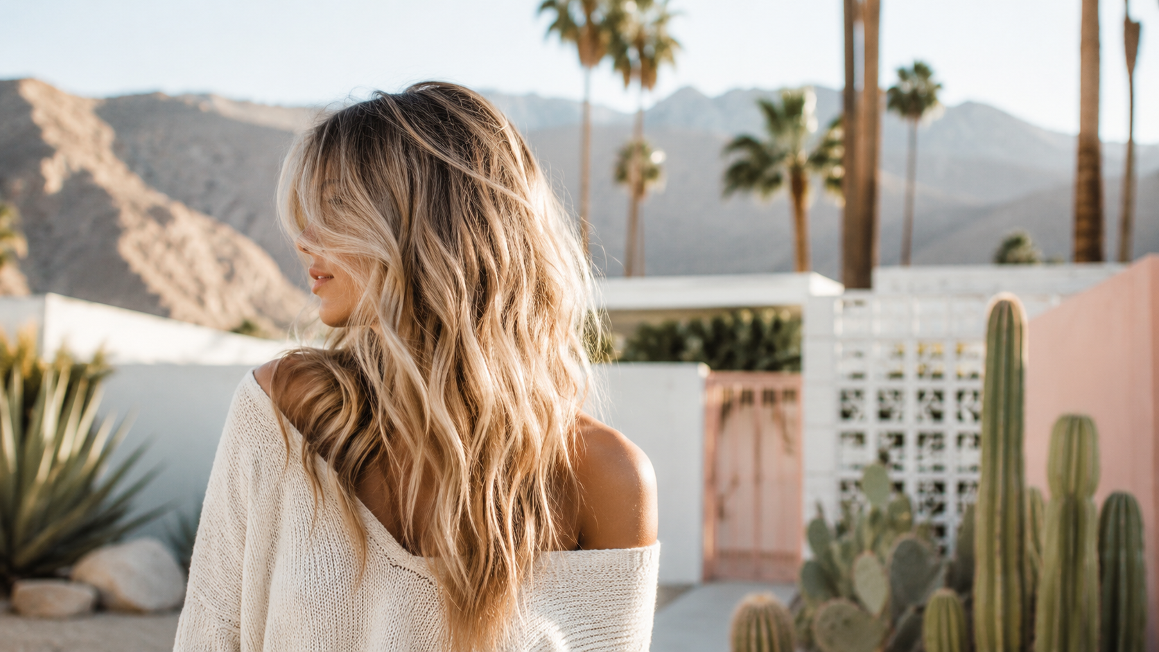 Woman with long wavy blonde hair in a white off-shoulder sweater standing outdoors in a desert landscape with mountains, palm trees, cacti, and colorful buildings in the background.