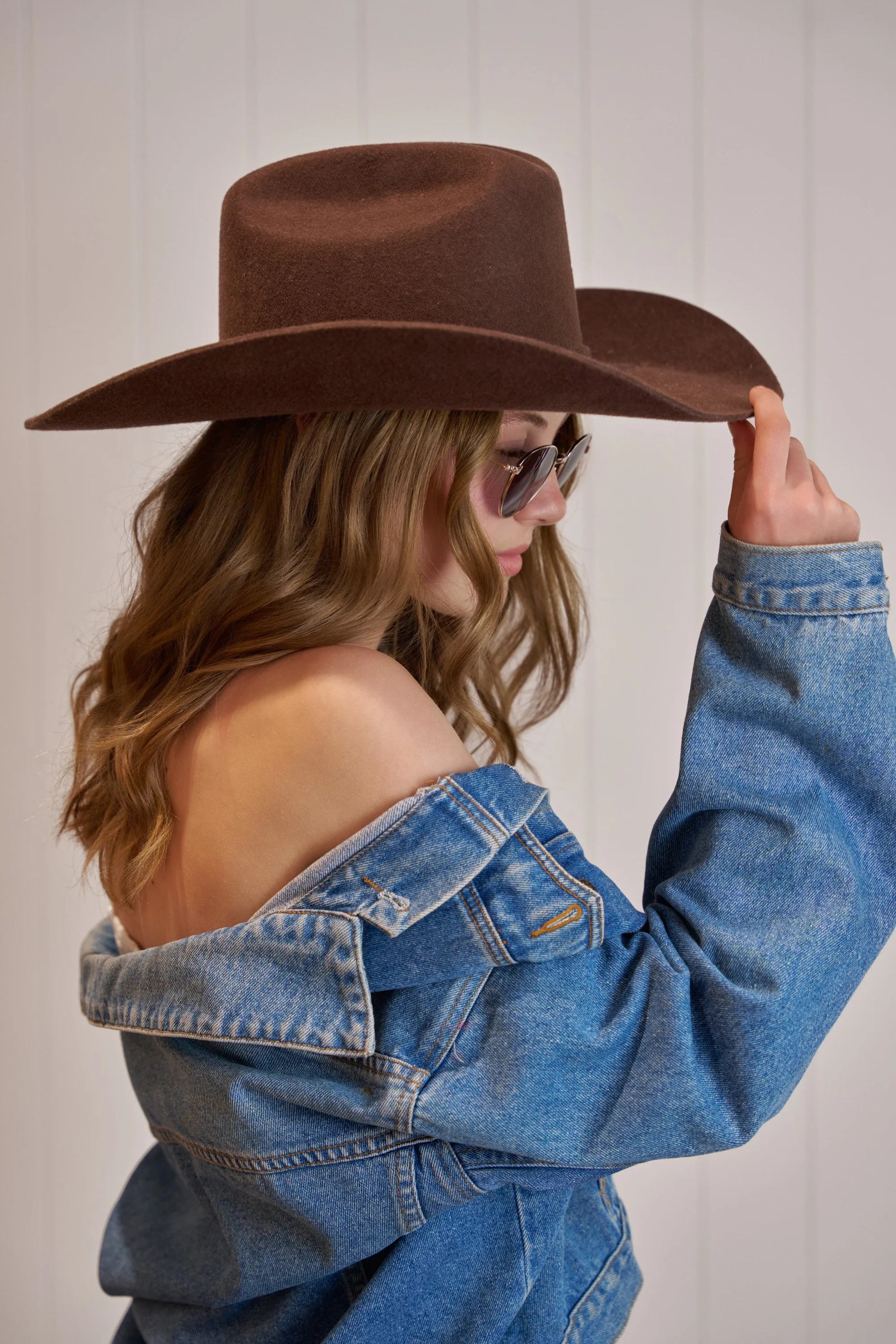 A woman with wavy brown hair wearing sunglasses, a denim jacket, and a large brown wide-brimmed hat touching the brim of the hat with her right hand.