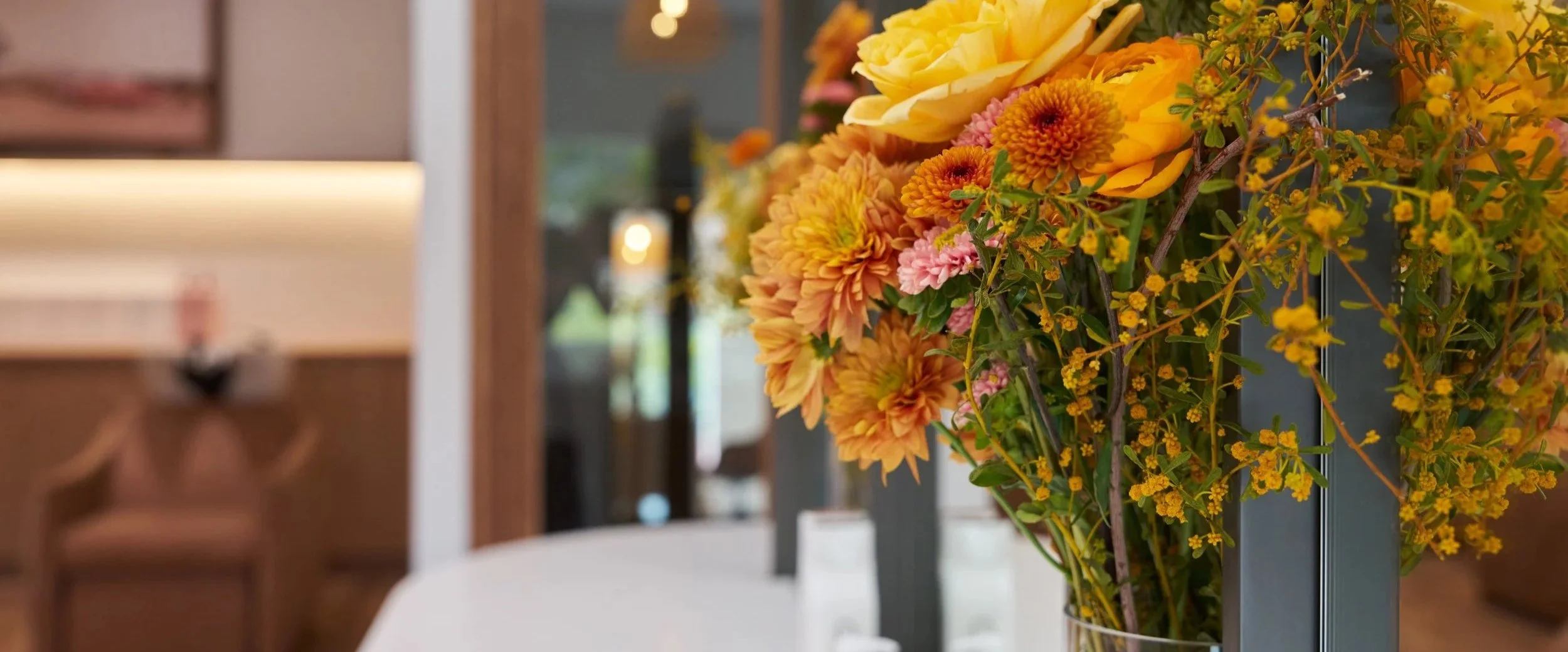 Close-up of a bouquet of yellow, orange, and pink flowers in a vase on a table, with a blurred indoor background.