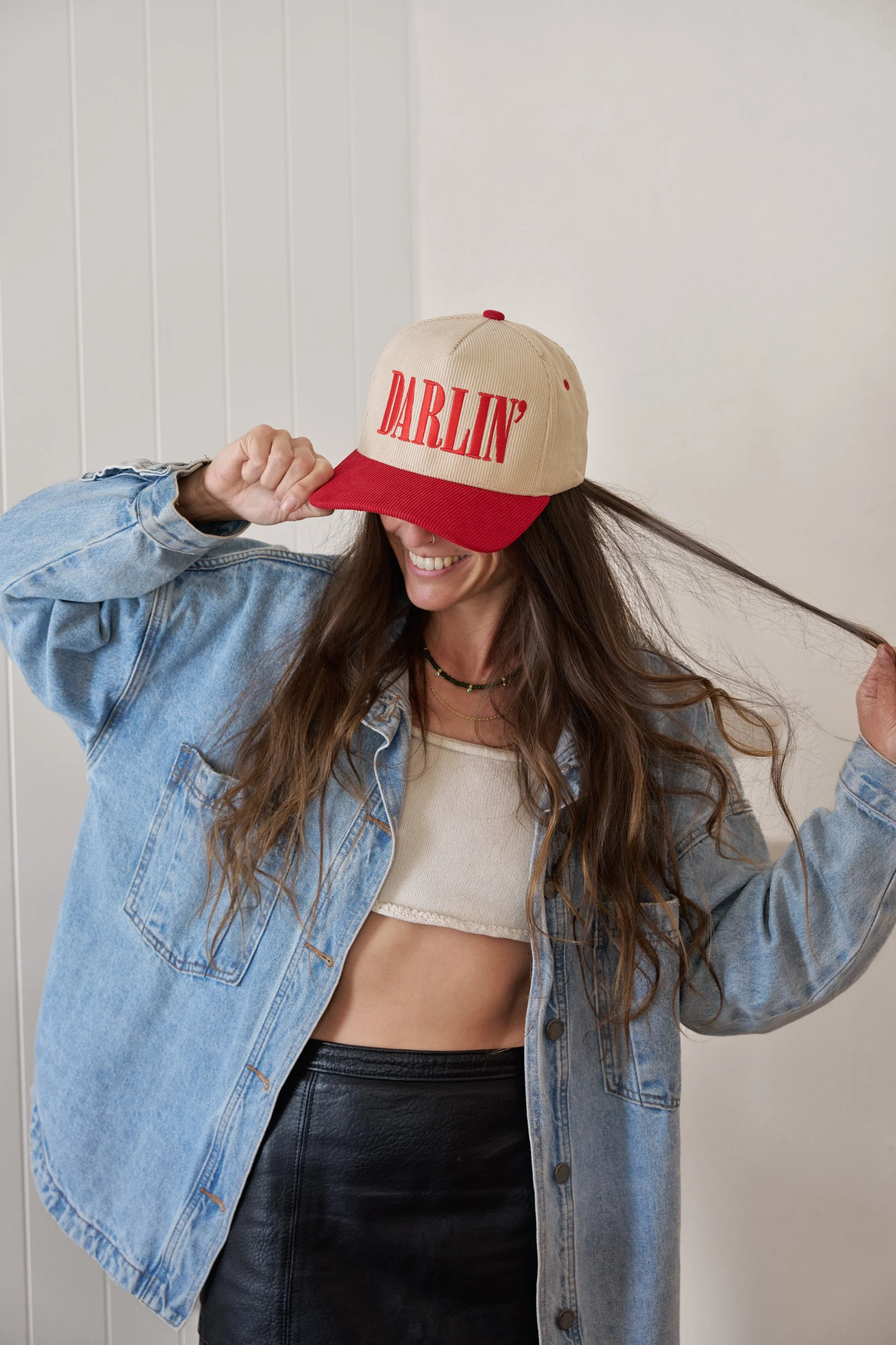 A woman with long, wavy brown hair wearing a beige and red cap with the word 'DARLIN' embroidered on it, a denim jacket, a beige crop top, and a black leather skirt, smiling, against a plain white wall background.