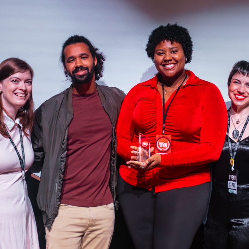 Four people standing together, one woman in the middle holding an award, smiling, at an event.