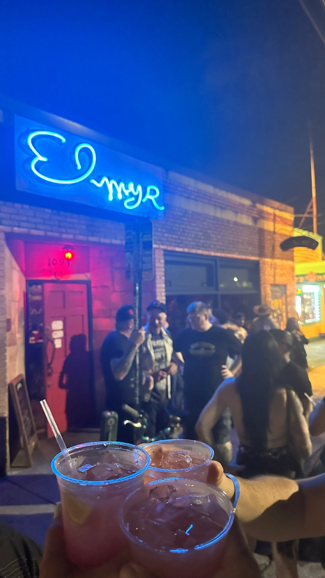 Night scene outside a bar or club with people socializing. Neon sign above reads 'Emy R' with a stylized bird logo. Two drinks with straws are held in the foreground.