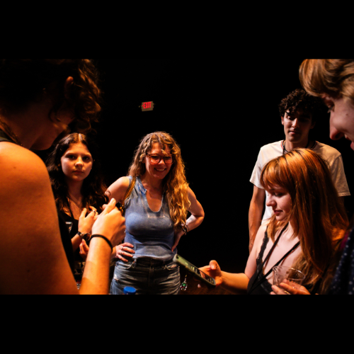 Group of five young women and girls gathered closely in conversation, smiling and engaging with each other in a dark room with dim lighting.