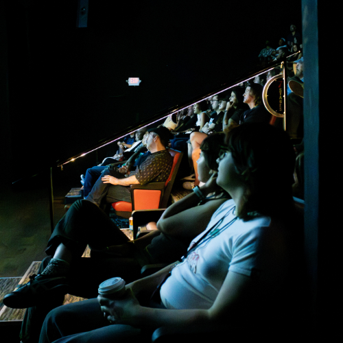 People sitting in a dark theater or auditorium watching a performance or presentation.