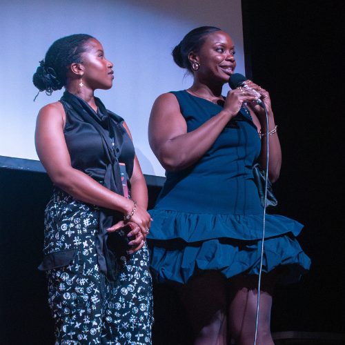 Two women standing on stage, one speaking into a microphone while the other listens, both dressed in formal black attire.