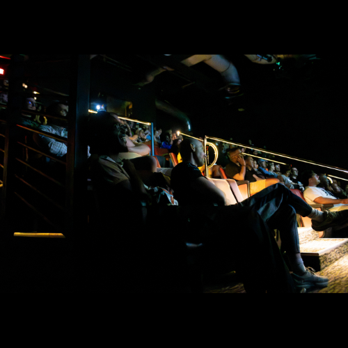 Audience members sitting in a dark theater or cinema with illuminated seats and a few lights on.