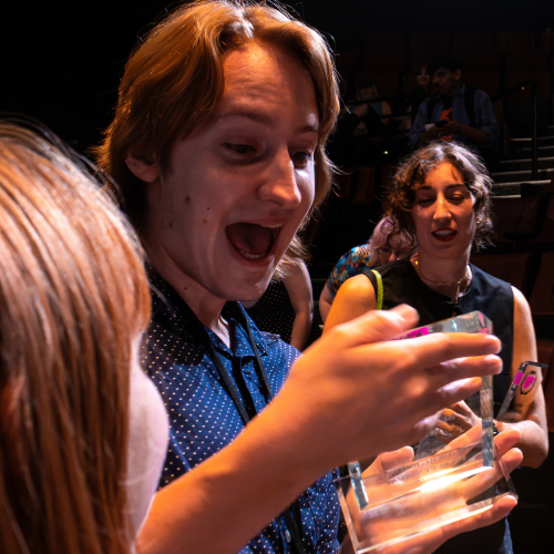 A young man with red hair and a blue shirt excitedly looking at a clear plastic box held by a girl with red hair. Two other women are in the background, one with curly hair holding a phone and another with dark hair and a black top, in a dimly lit indoor setting.