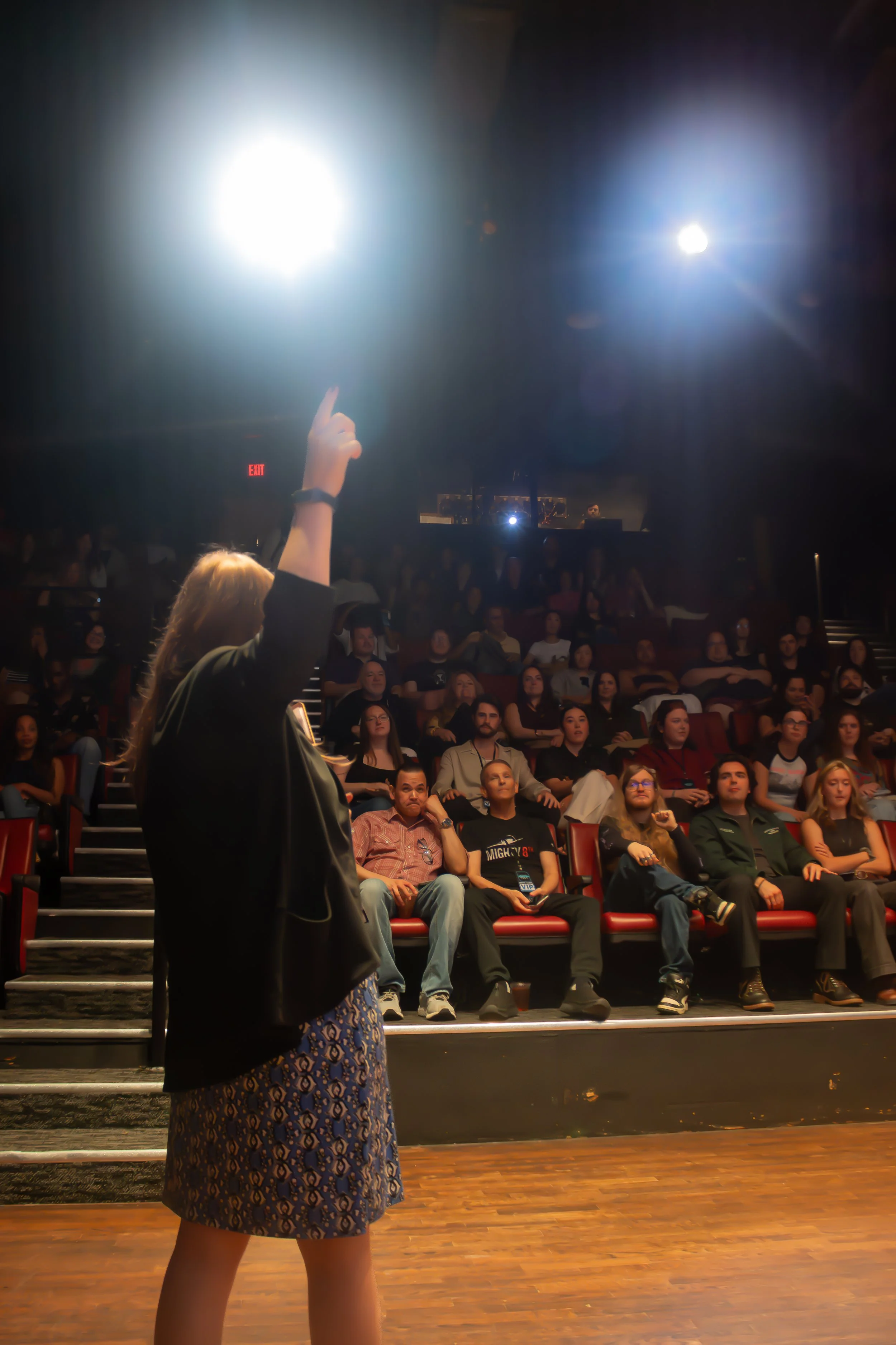 A woman with long hair and wearing a patterned skirt and a black jacket gestures toward bright lights on stage in front of an audience seated in a theater with red seats and dark walls.