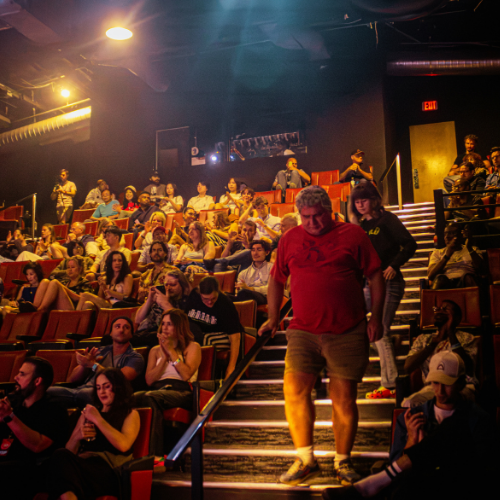 Audience members in a theater or auditorium, some seated and some descending stairs, with warm lighting and a dark background.
