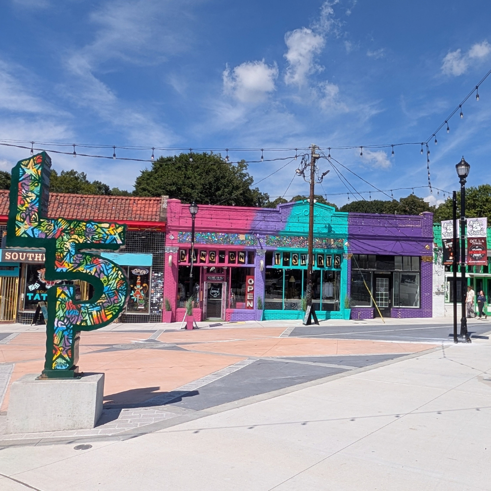 Colorful street scene with storefronts painted in pink, purple, and turquoise, a large decorative letter 'S' sculpture, and string lights overhead.