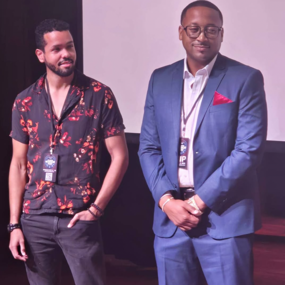 Two men standing next to each other, one wearing a floral shirt and the other in a blue suit, both with event badges around their necks indoors.