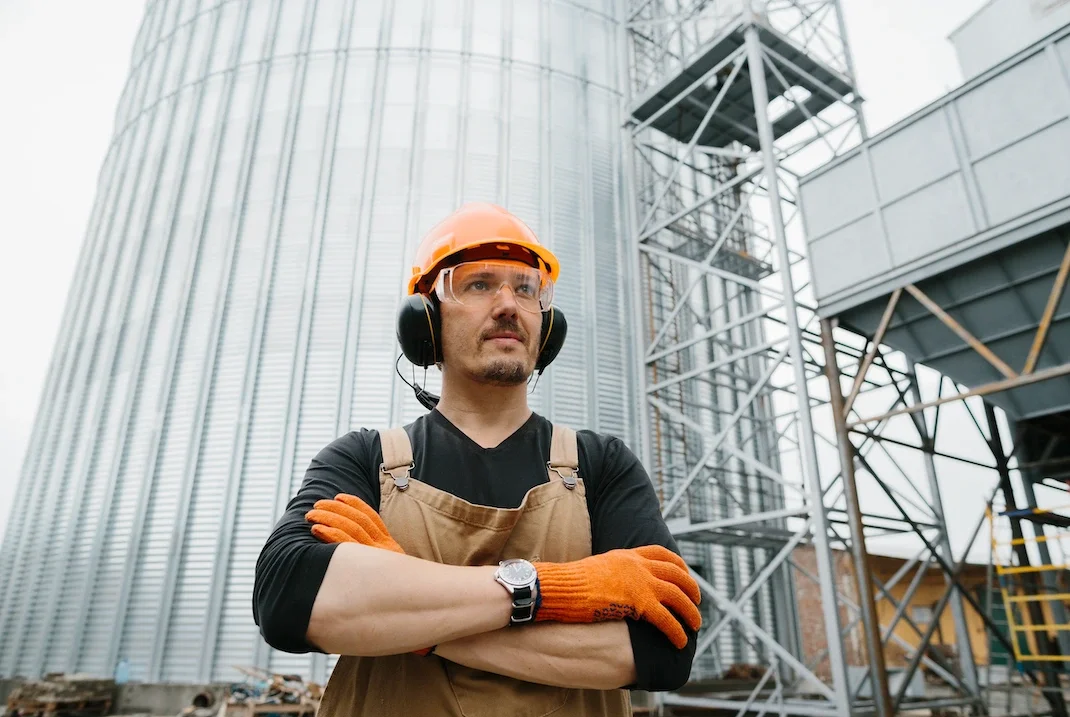 Construction worker with safety helmet, goggles, and orange gloves standing with arms crossed in front of large industrial storage silos and scaffolding.