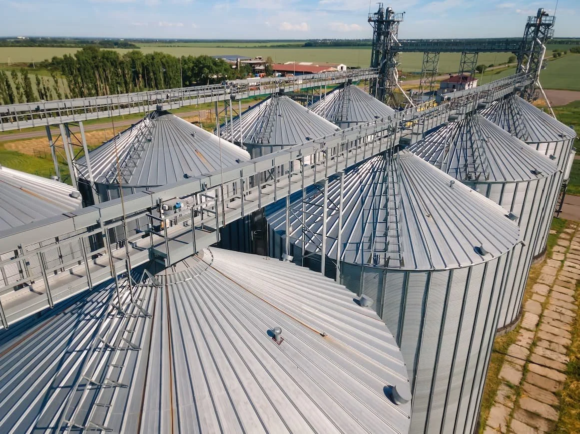 Aerial view of a grain silo storage facility with multiple metal silos and connecting conveyor systems in a rural area.