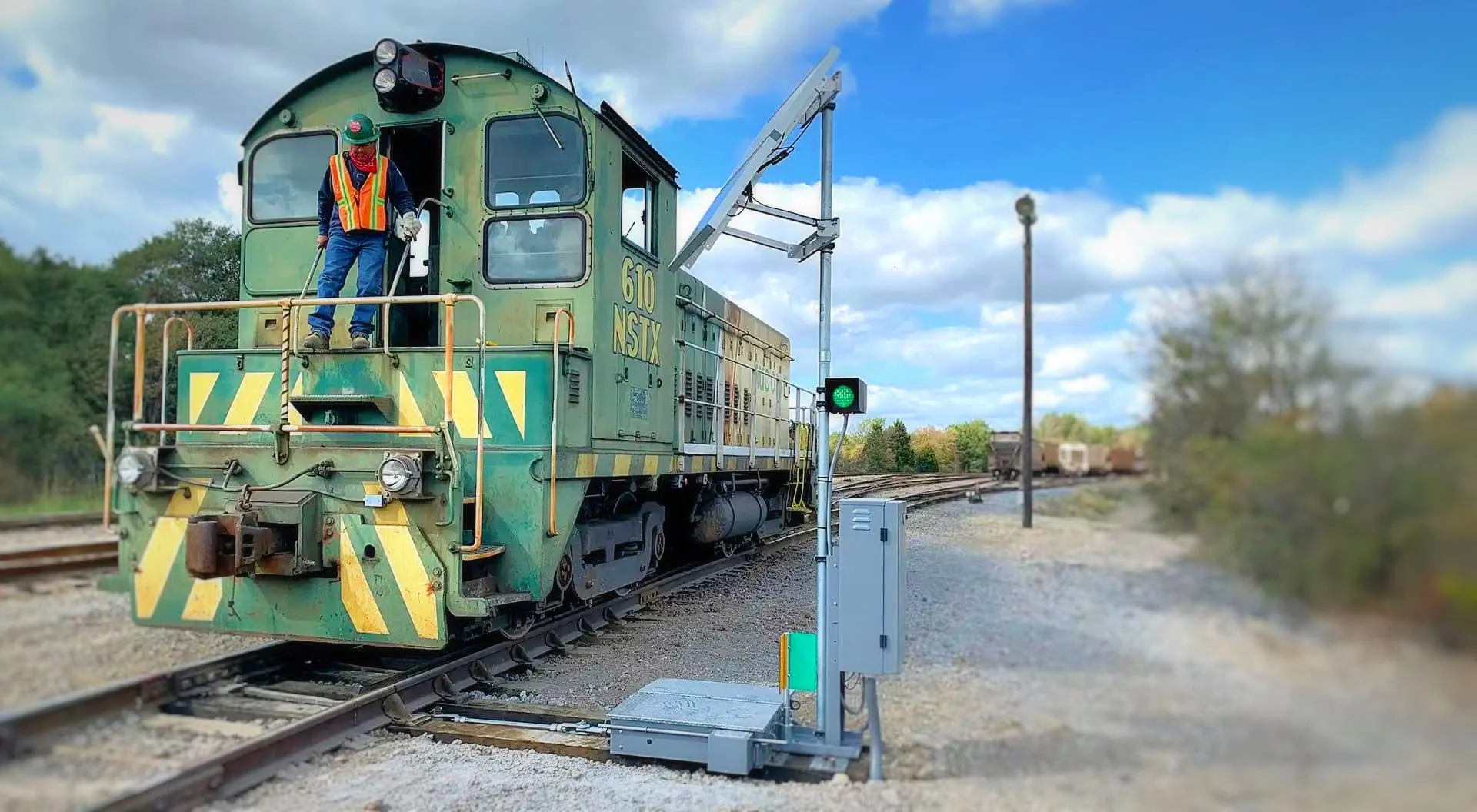 A green rail maintenance locomotive with a worker in a safety vest and helmet on a railroad track, near a signaling device showing a green light, with additional railroad cars in the distance under a partly cloudy sky.