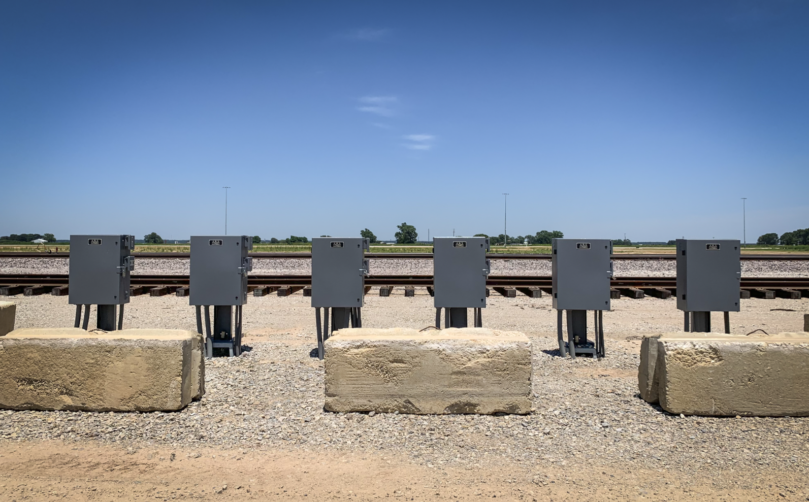 Six gray utility boxes mounted on short metal poles along train tracks in an open area with a clear blue sky and a few distant trees.
