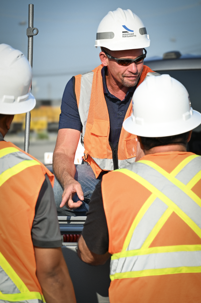 Three construction workers wearing hard hats and safety vests having a discussion outdoors at a construction site.