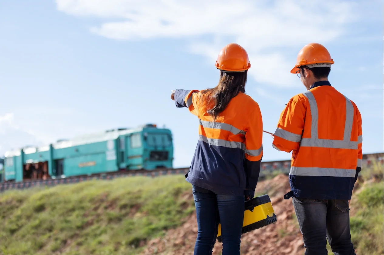 Two construction workers in orange safety vests and helmets standing outdoors near train tracks, one pointing towards a moving train with a blue sky background.