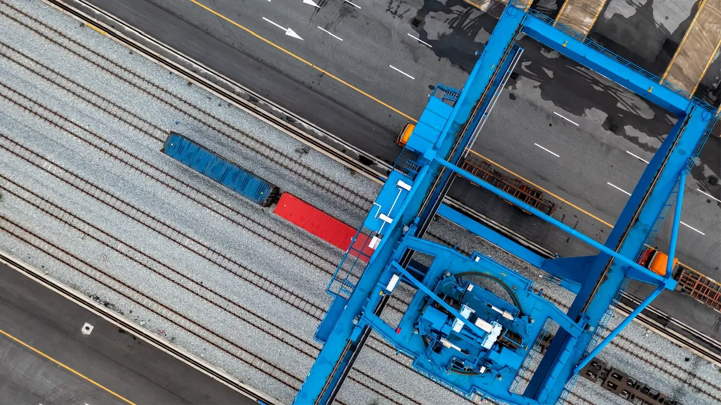 Aerial view of a blue crane lifting freight containers on railway tracks.