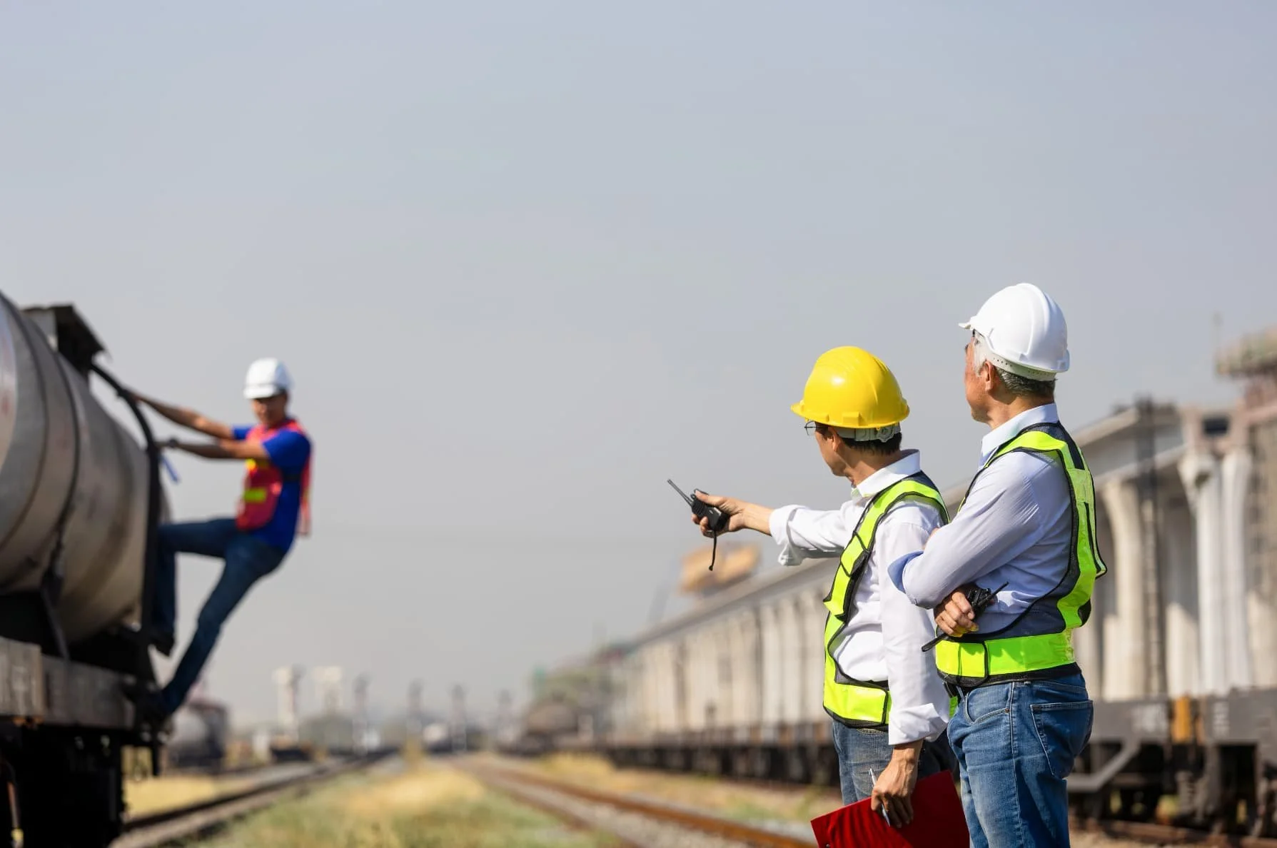 Two construction engineers with yellow and white safety helmets and high-visibility vests observing and communicating near railway tracks, while a worker in a white helmet and high-visibility vest works on a pipeline or train component in the background.