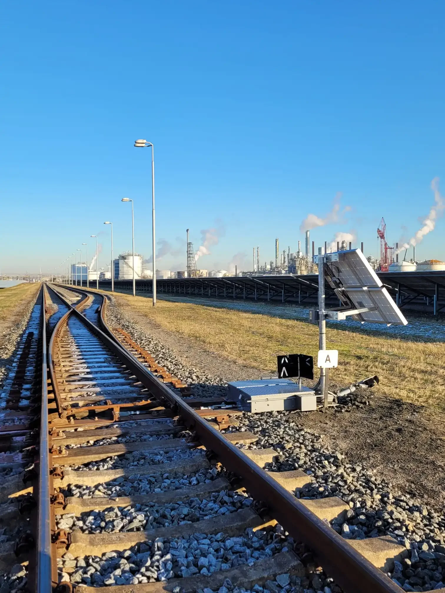A solar panel beside railroad tracks in an industrial area with factories emitting smoke in the background.