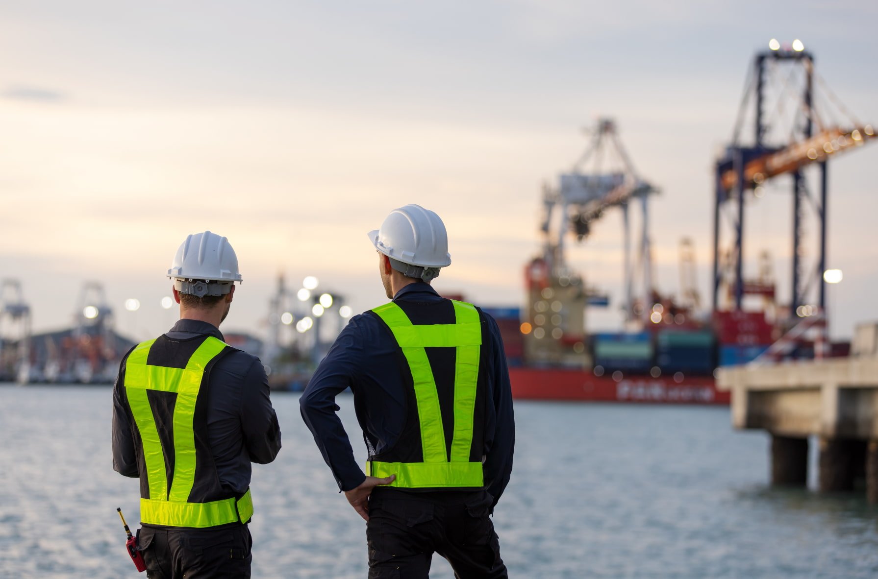 Two men wearing white safety helmets and reflective vests standing by the water, looking at industrial cranes and cargo ships at a port during sunset.