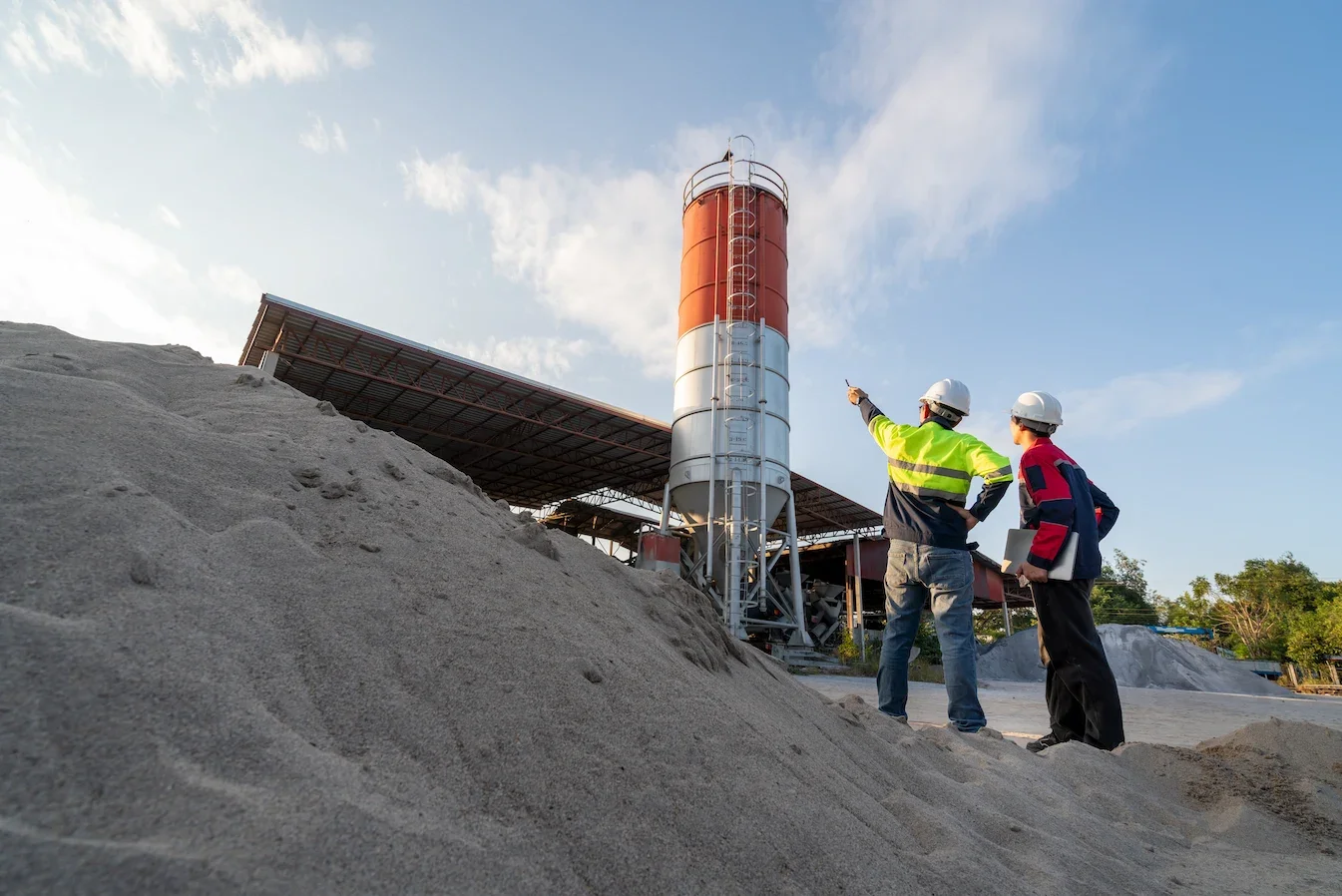 Two construction workers wearing safety helmets and reflective jackets inspecting a large industrial silo or tower at a construction site.