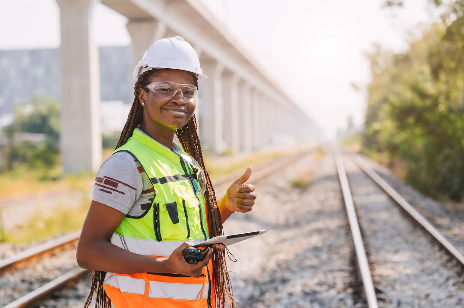 A woman wearing a white safety helmet, safety glasses, and a high-visibility vest standing next to railroad tracks under a bridge, giving a thumbs up and holding a tablet in her hand.