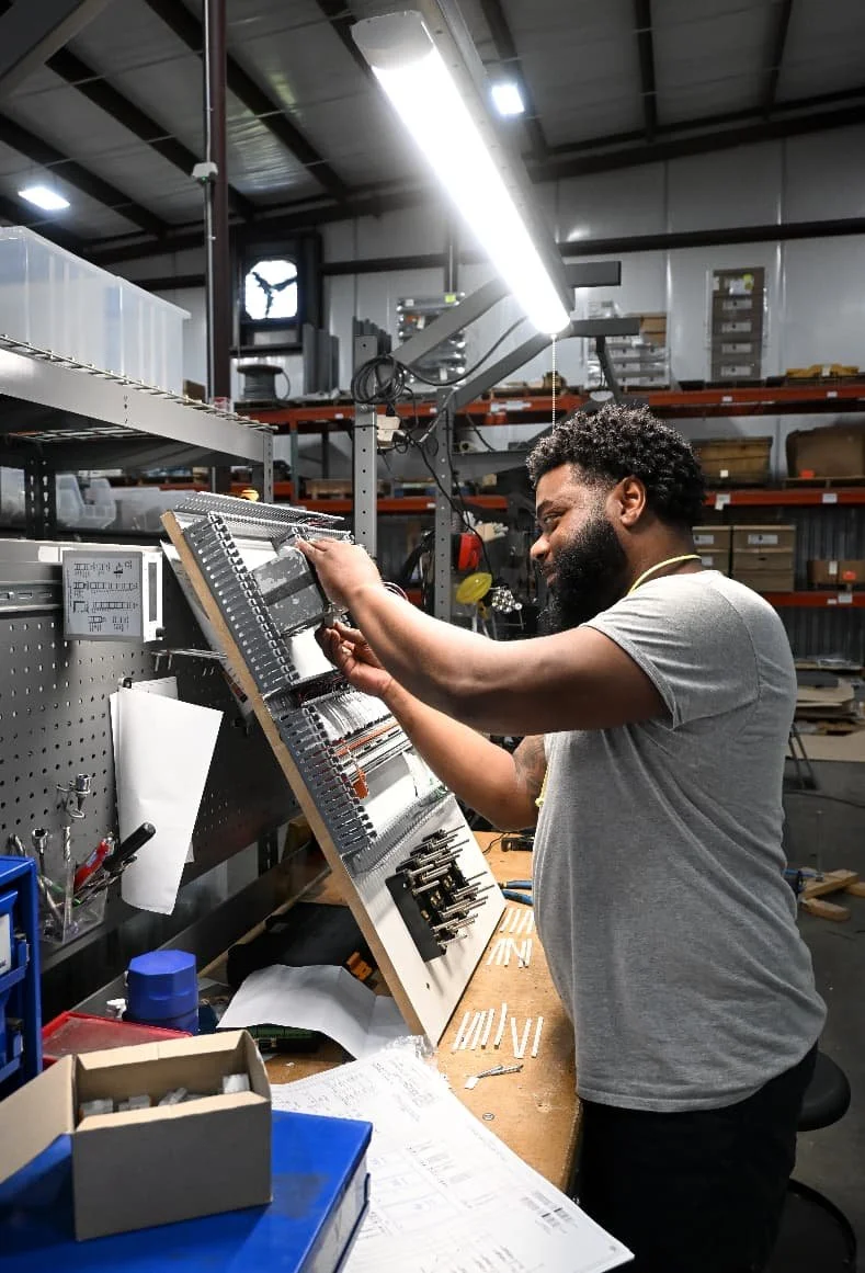 A man working on an electronic circuit board in a warehouse or workshop, with shelves and tools around him.