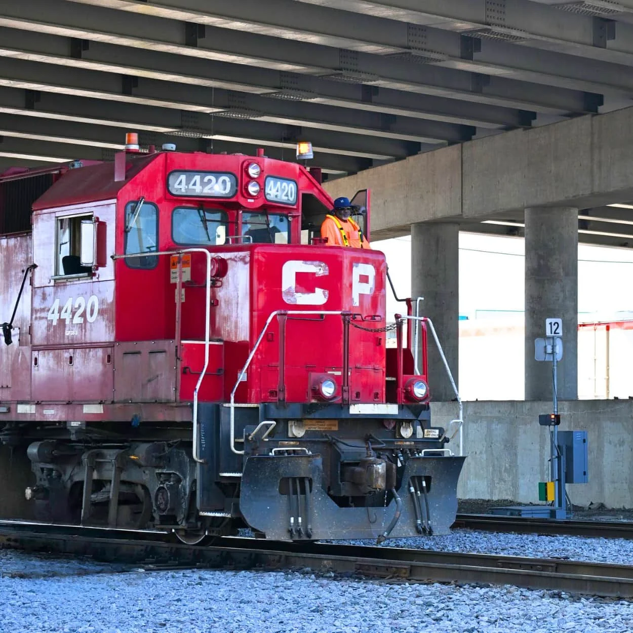 A red Canadian Pacific train locomotive numbered 4420, with a worker in orange safety gear and a blue cap, standing on a rail track under a concrete bridge.