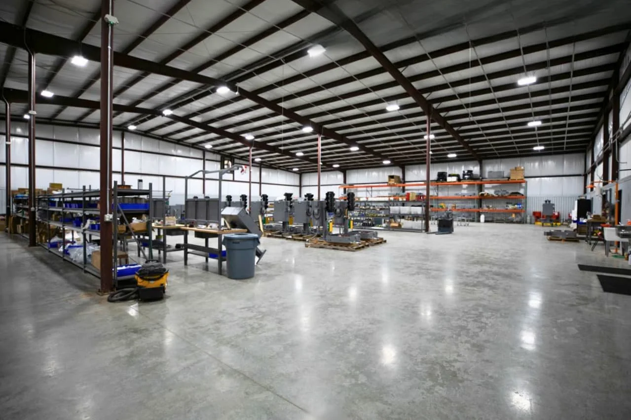 Empty industrial warehouse with metallic shelves, workbenches, and machinery, well-lit with overhead lights and a concrete floor.