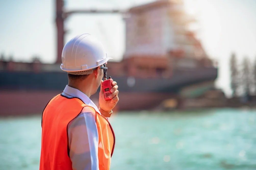 A person wearing a white safety helmet, orange safety vest, and a light-colored shirt, speaking into a red handheld radio near a body of water with a large ship in the background.