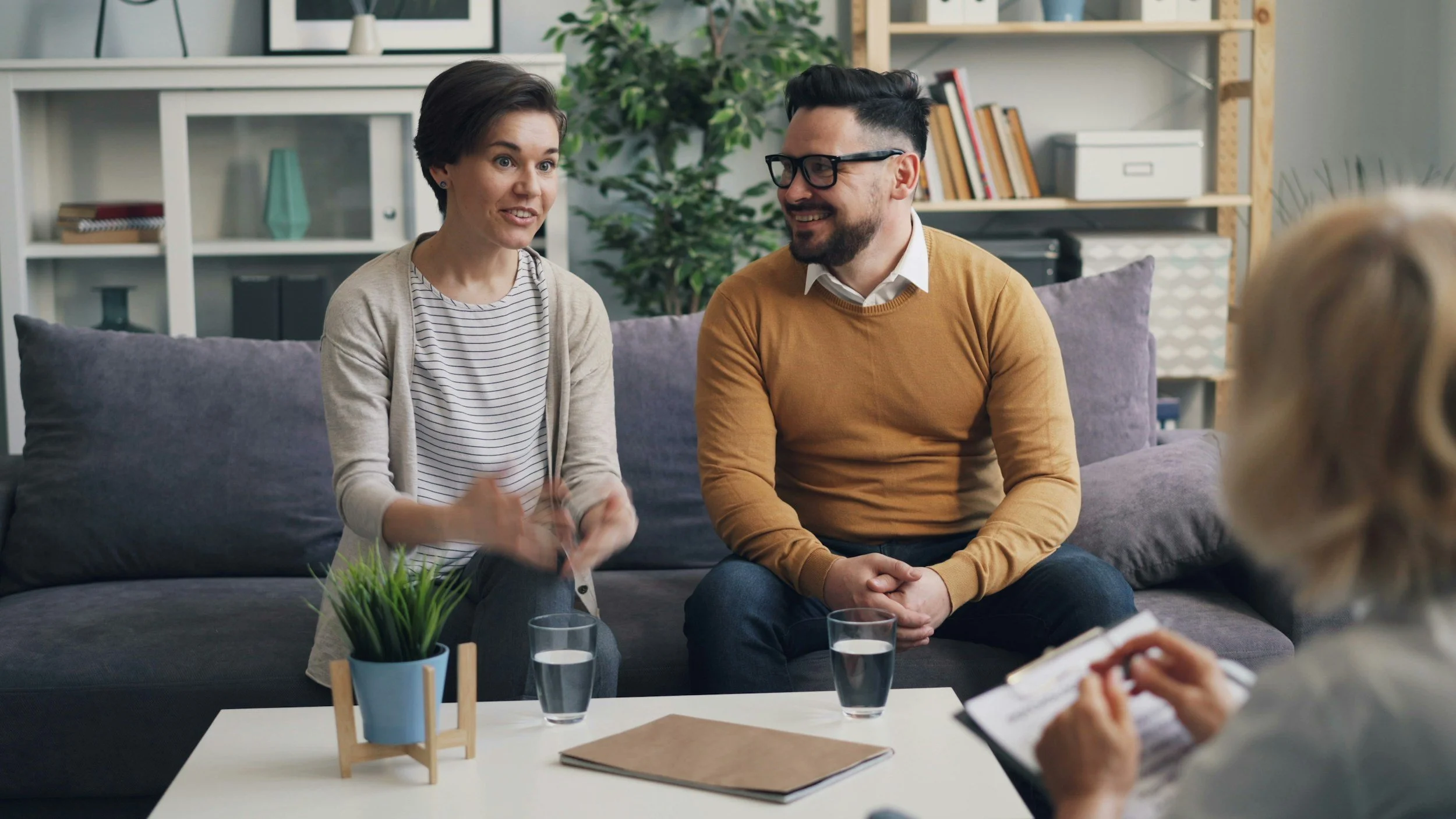 Two people sitting on a sofa in a living room, engaged in conversation. A woman with short dark hair wearing a striped shirt and a beige cardigan is speaking, while a man with glasses and a beard in a mustard sweater listens, smiling. In the foreground, a white table holds glasses of water, a potted plant, and notebooks.