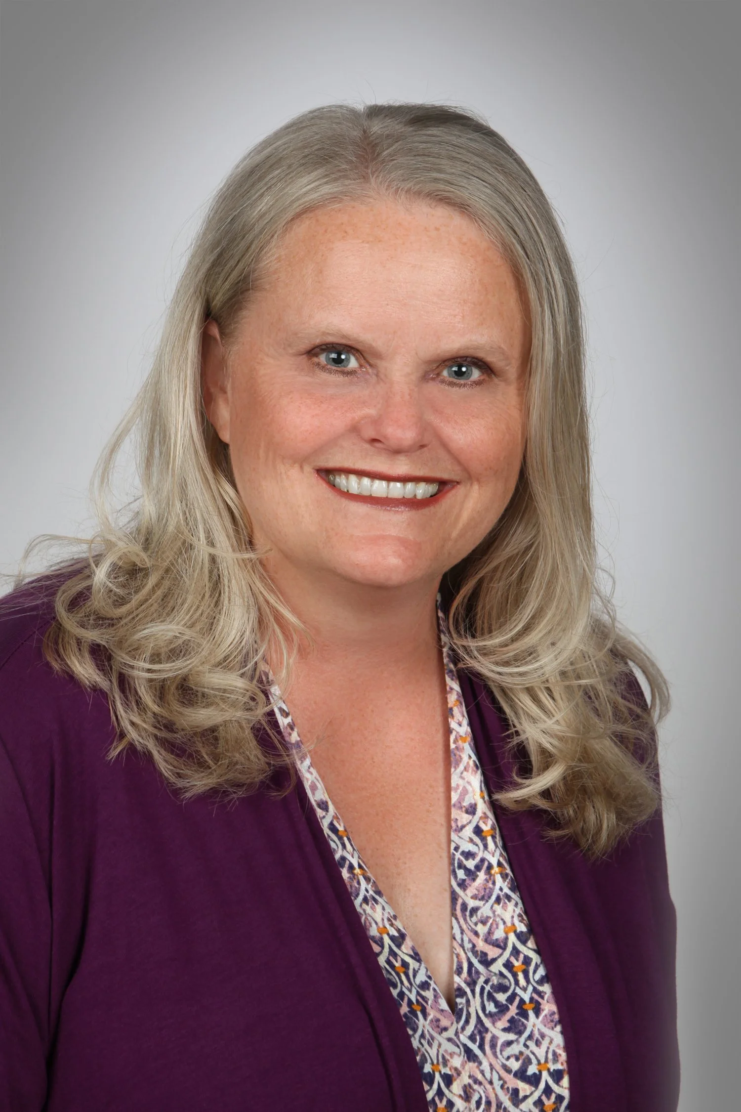 A woman with long, curly blonde hair, blue eyes, and a broad smile, wearing a purple blazer over a patterned blouse, against a plain, light gray background.
