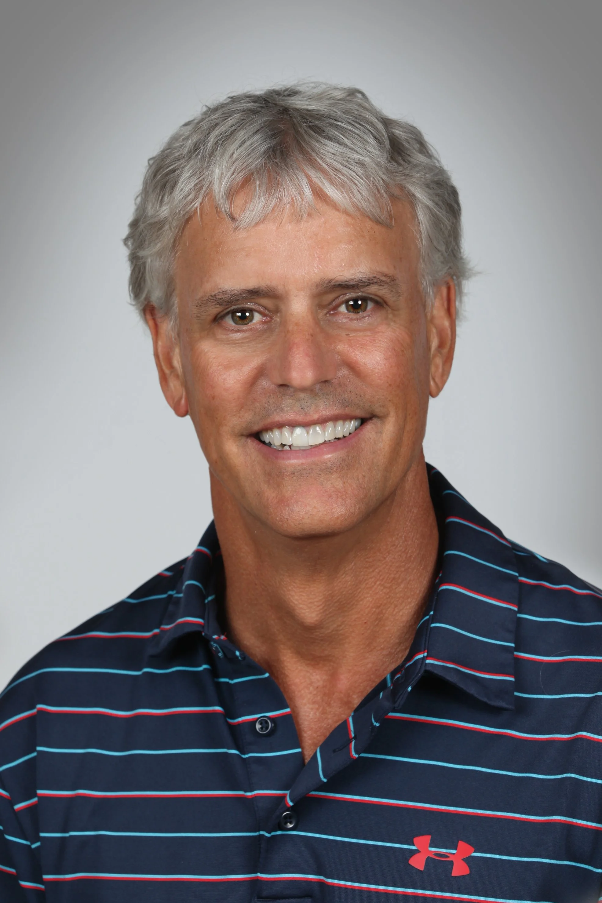 A smiling middle-aged man with gray hair wearing a navy blue polo shirt with red and white horizontal stripes and a red Under Armour logo.