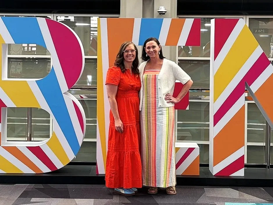Two women standing in front of large, colorful block letters spelling out "BLI" at an indoor event.