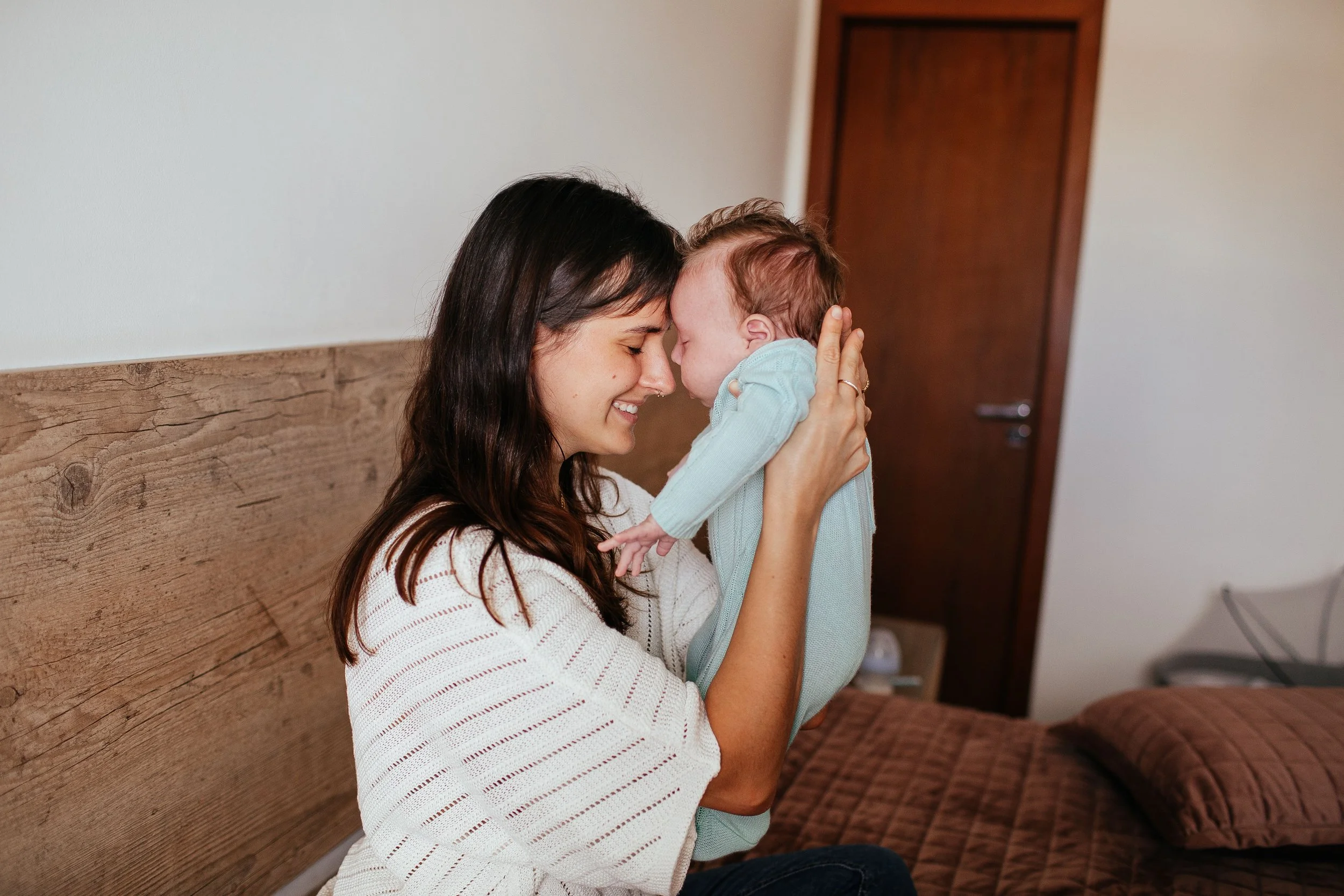 A woman is holding a baby close to her face, both with eyes closed, touching foreheads in an intimate moment in a warmly lit room.