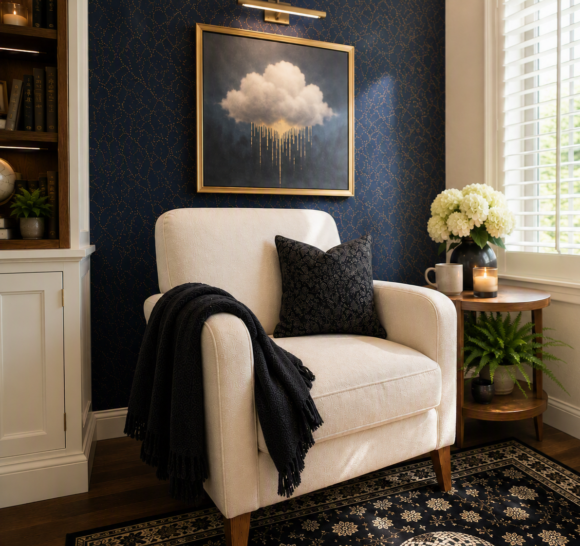 A cozy living room corner with a white armchair, black pillow, black throw blanket, a side table with a potted fern, white hydrangea flowers, candle, mug, and a framed cloud art painting on a dark patterned wall during the daytime.
